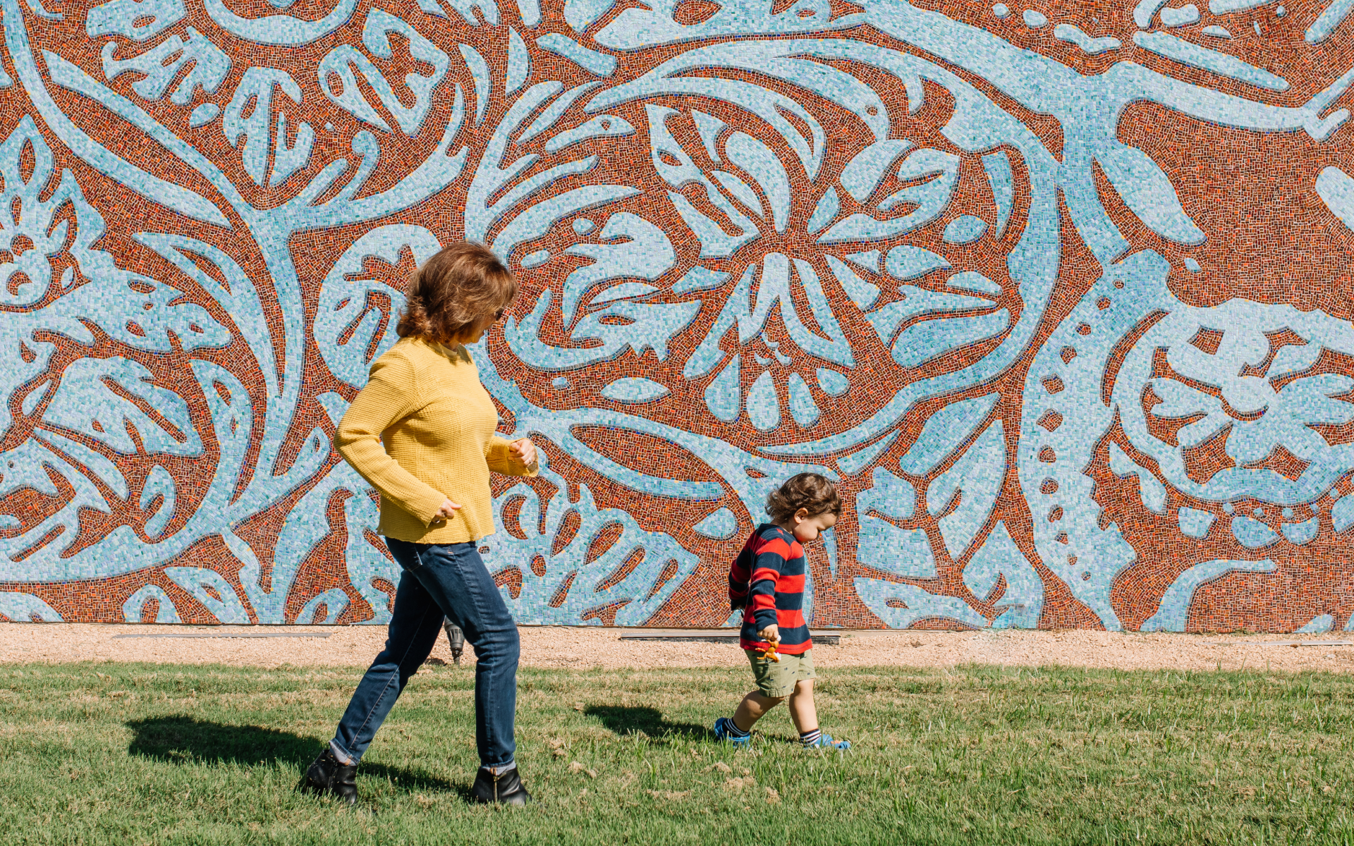 A woman in a yellow sweater and a child in a striped shirt walk on grass in front of a large, blue-and-red floral patterned wall.