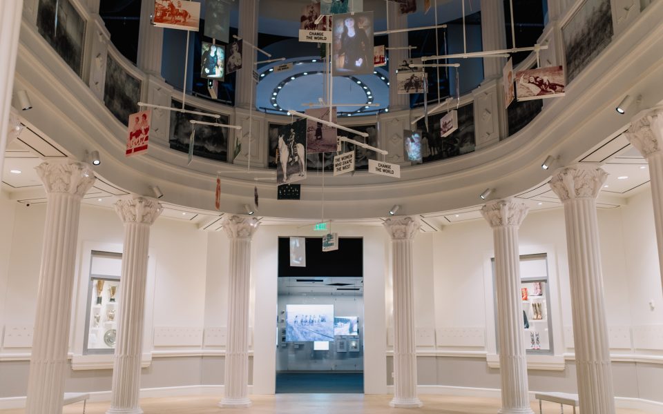 A museum exhibit with white columns, a circular balcony above, and various photos and signs suspended from the ceiling. The room is spacious, well-lit, and displays items and images on the walls and in the background.