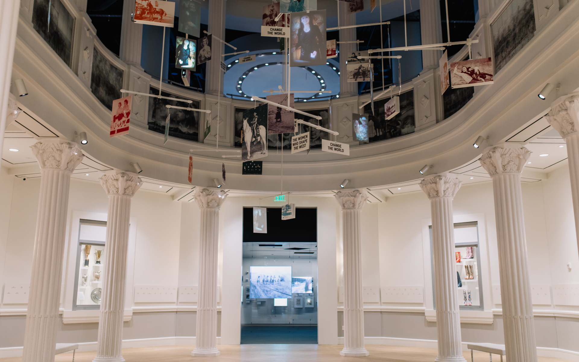 A museum exhibit with white columns, a circular balcony above, and various photos and signs suspended from the ceiling. The room is spacious, well-lit, and displays items and images on the walls and in the background.