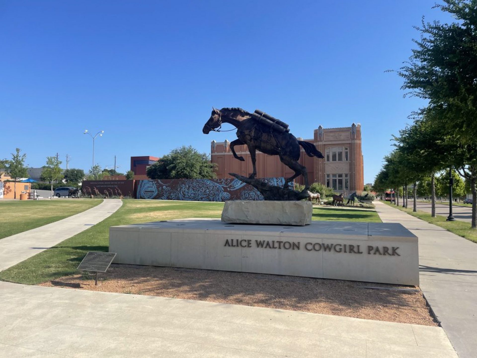 A bronze statue of a cowgirl riding a horse stands on a stone pedestal labeled Alice Walton Cowgirl Park, with a historic brick building and green lawn in the background under a clear blue sky.
