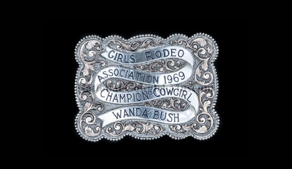 An ornate silver belt buckle reads Girls Rodeo Association 1969 Champion Cowgirl Wanda Bush with decorative scrollwork on a black background.