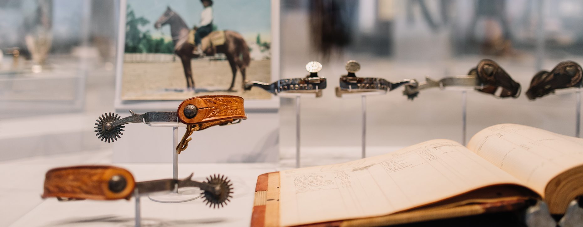 Several vintage multi-tools and a leather-bound book are displayed in a glass case, with a blurred photograph of a person riding a horse in the background.