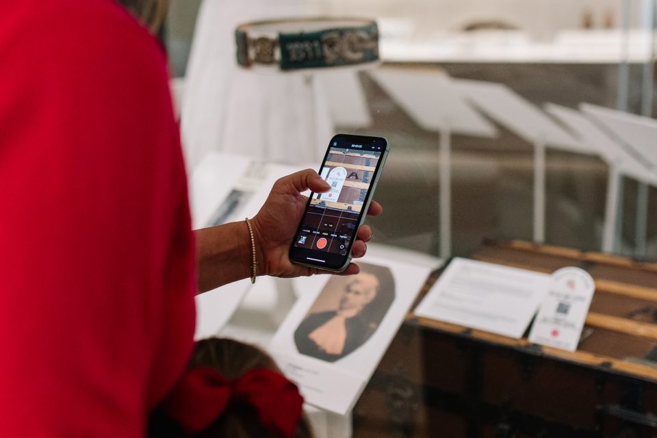 A person in a red outfit takes a photo of a museum exhibit with their smartphone; display cases and informational plaques are visible in the background.