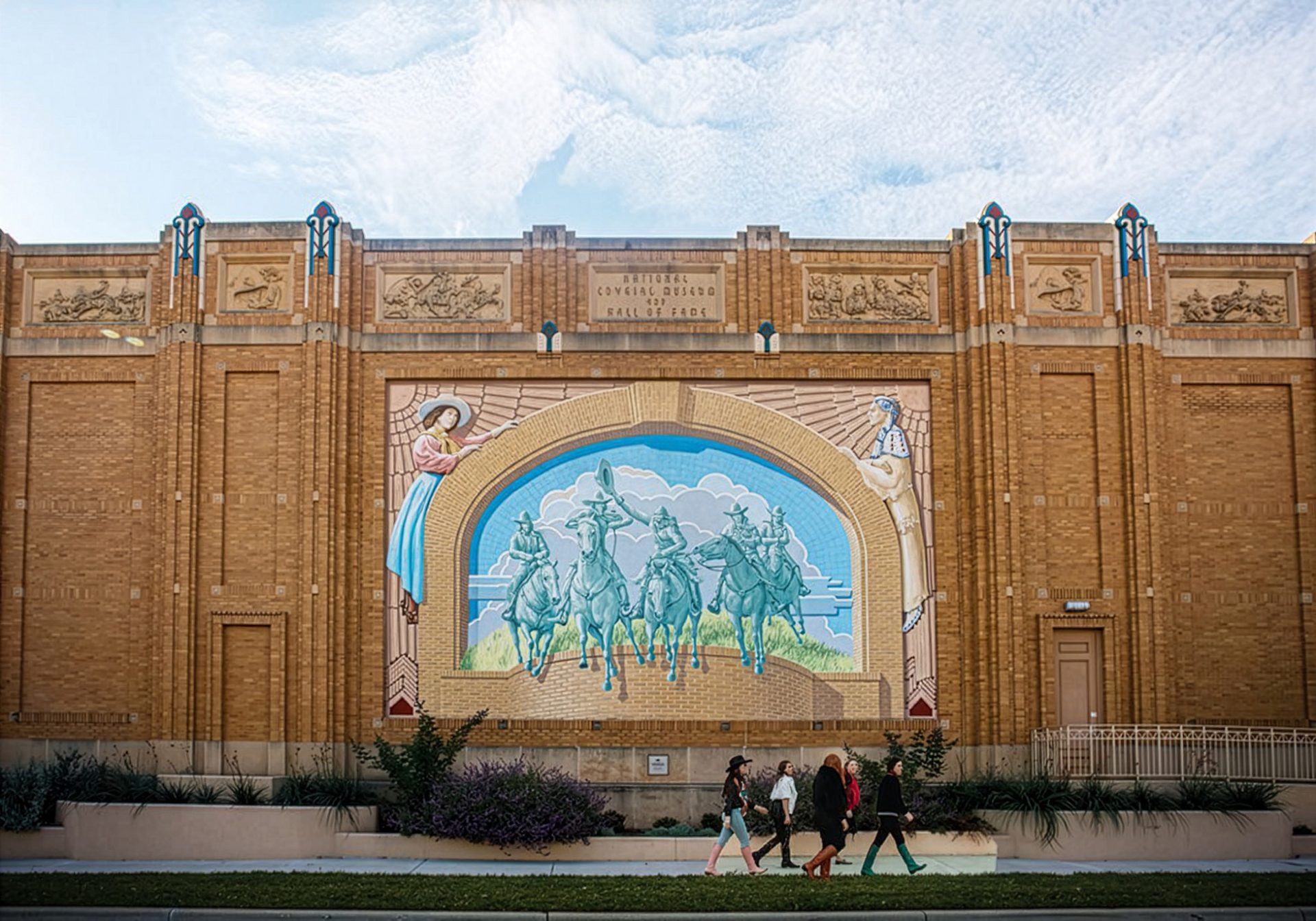 A large mural of five horseback riders decorates a brick building as four people walk by on the sidewalk below, under a partly cloudy sky.