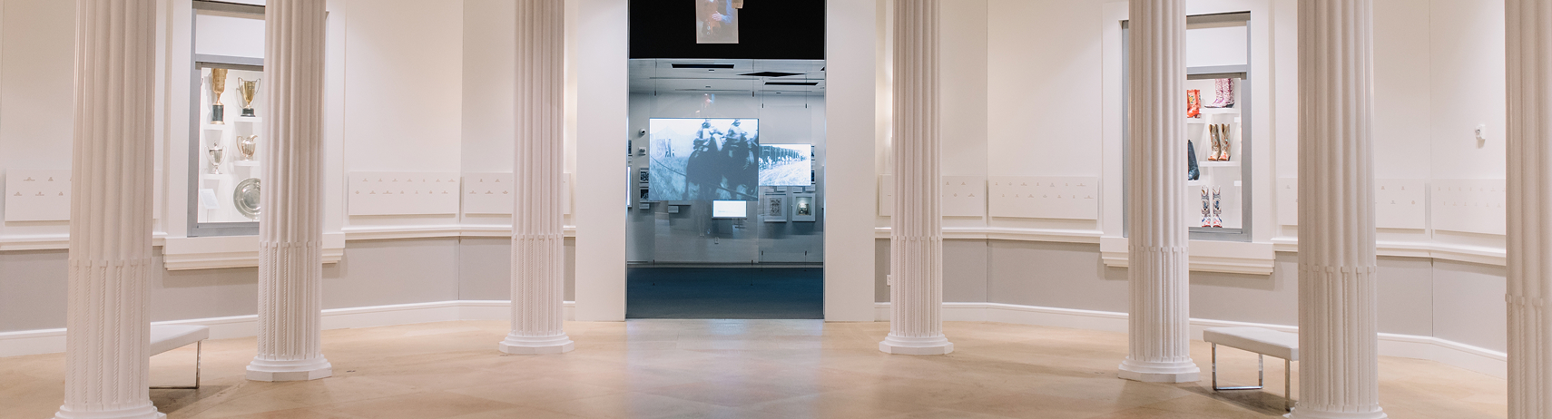 A bright museum gallery with white columns, beige floors, and exhibits in glass cases along the walls. A doorway in the center leads to a darker room with a large projected black-and-white image.