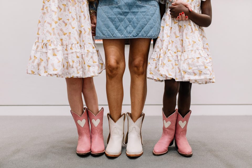 Three people stand side by side. Two children in matching patterned dresses and pink cowboy boots with white hearts, and an adult in a blue dress and white cowboy boots. Only their legs are visible.