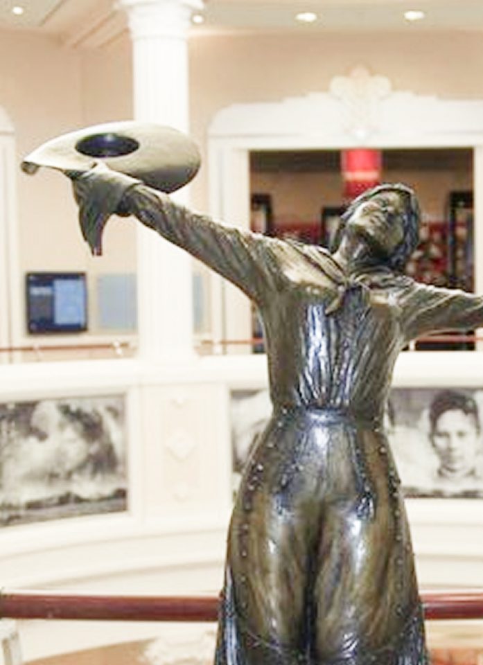 A bronze statue of a woman in old-fashioned attire holds a hat aloft with one hand, appearing joyful. The background includes portraits and museum decor.