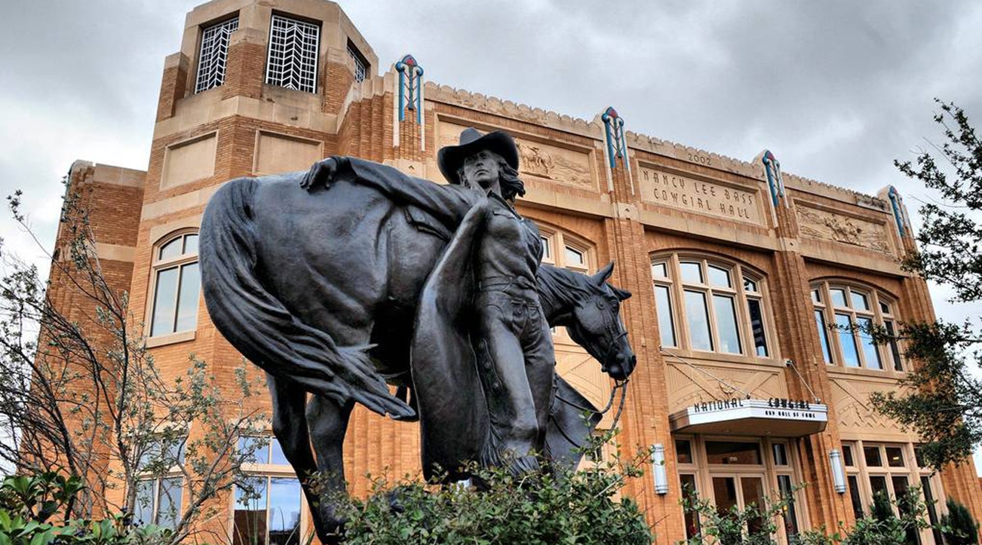 A bronze statue of a cowboy and horse stands in front of a historic brick building with decorative windows and FORT WORTH STOCKYARDS on the facade; trees and cloudy sky in the background.