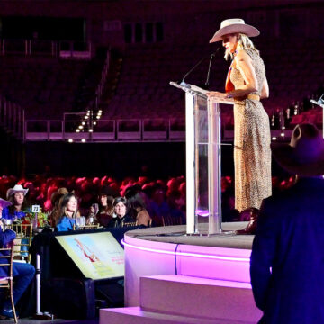 Female speaker in a leopard-print dress and pink cowboy hat speaks at a clear podium on a lit stage, addressing an audience in cowboy hats.