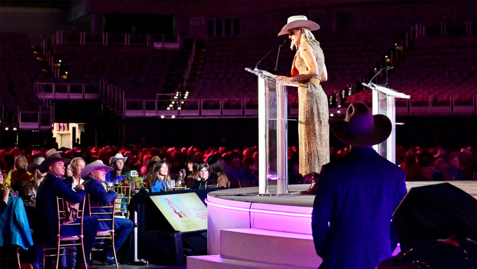 Female speaker in a leopard-print dress and pink cowboy hat speaks at a clear podium on a lit stage, addressing an audience in cowboy hats.