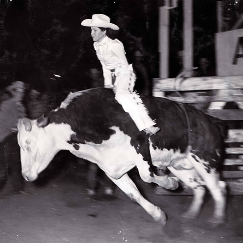 A rodeo rider in a white outfit and cowboy hat rides a bucking bull at night, surrounded by blurred spectators and arena fencing.