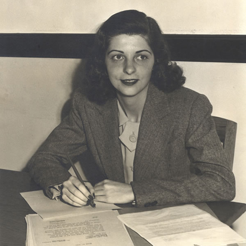 A woman with dark hair wearing a suit jacket sits at a desk, holding a pencil, and looks slightly to the side. Papers are spread out on the desk in front of her. The photo is black and white.