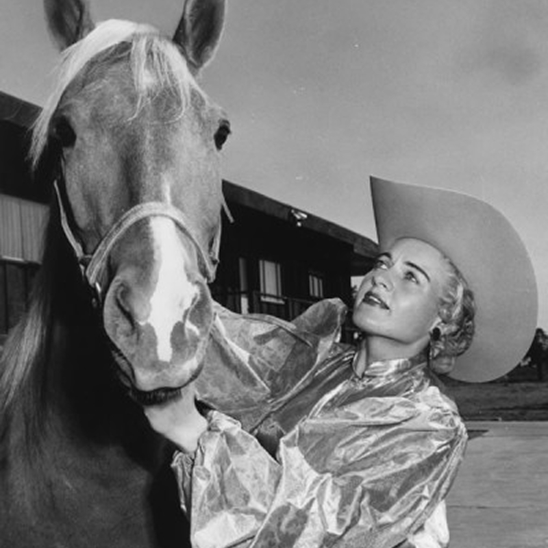 A woman wearing a shiny jacket and cowboy hat stands next to a horse, looking up and smiling. The background shows a building and some trees under a partly cloudy sky.