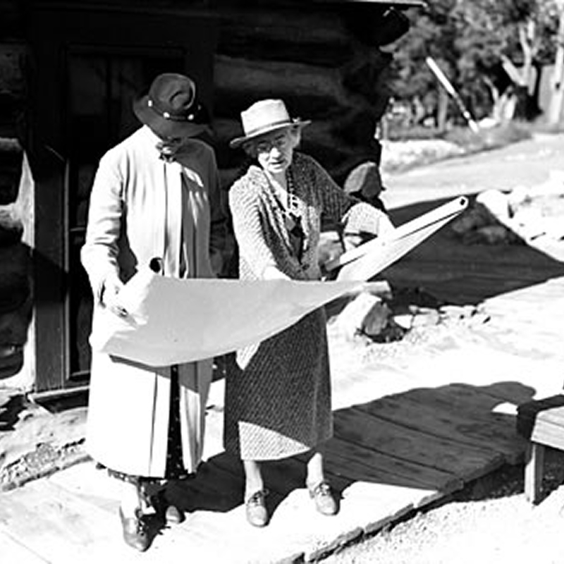 Two women in hats and coats stand outdoors, holding and examining a large sheet of paper together, possibly a blueprint or map, near a wooden building on a sunny day.