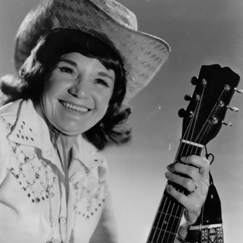 A woman wearing a cowboy hat and embroidered shirt smiles while holding a guitar, posed against a plain background.