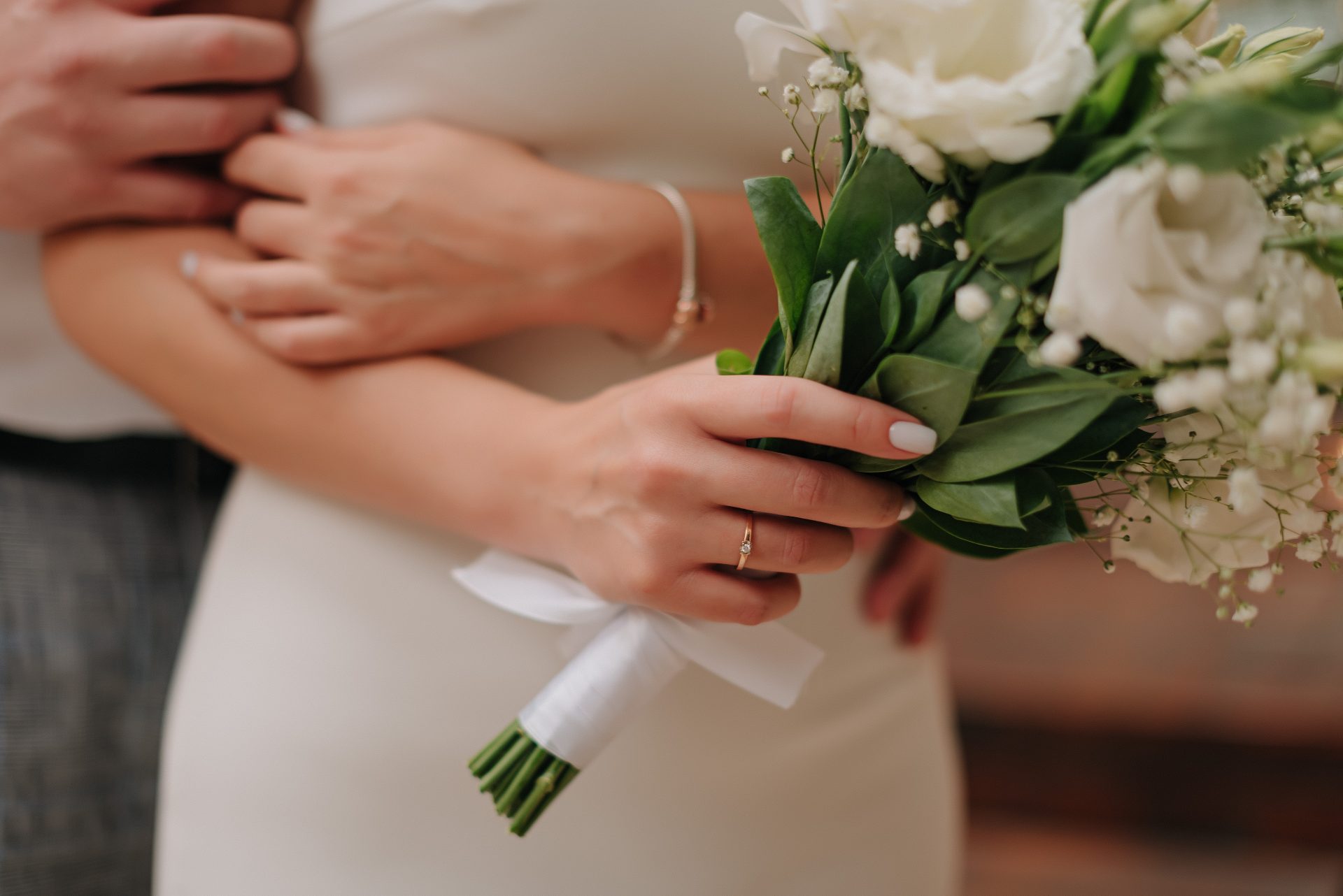 A bride holding a bouquet of white flowers is embraced by another person. The focus is on her hand, showing a ring and a simple bracelet, against the soft white of her dress.