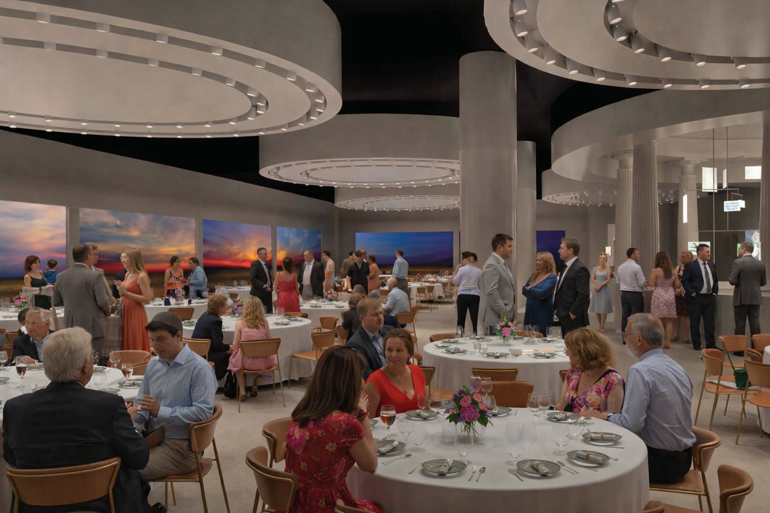 People in formal attire are seated at round tables and standing, mingling at an elegant indoor event with modern lighting and large wall art featuring sunset landscapes. Some tables have floral centerpieces.