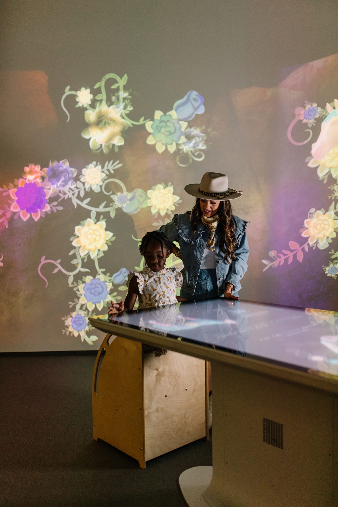 A woman and a child interact with a large touchscreen table, with colorful floral and bird projections on the wall behind them.