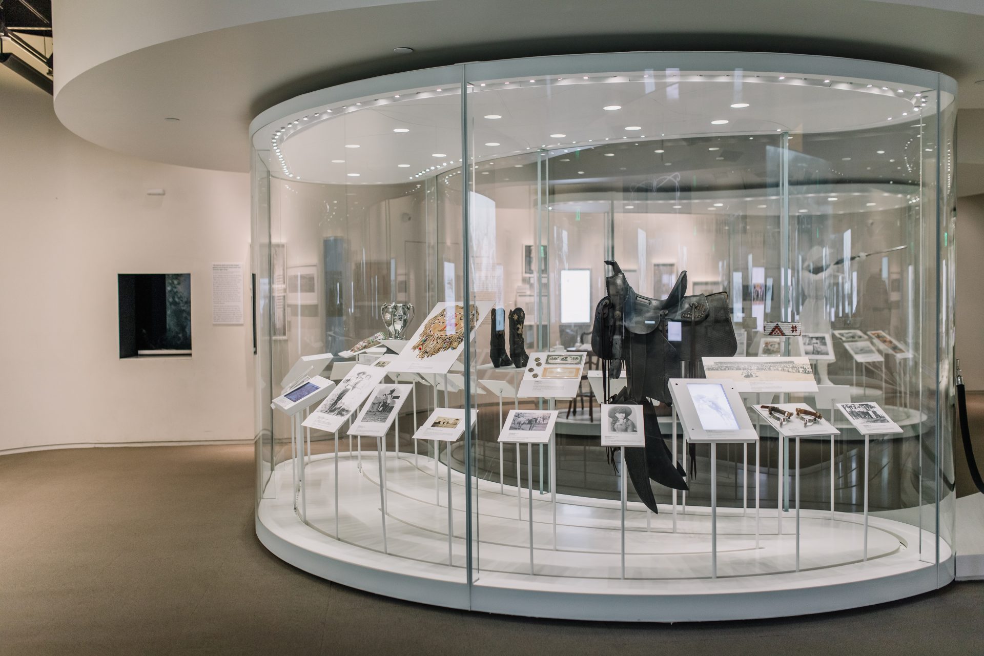 A glass display case in a museum exhibits historical artifacts, documents, and photographs. The case is well-lit, with descriptive placards and mounted items inside, set in a modern, white-walled gallery space.