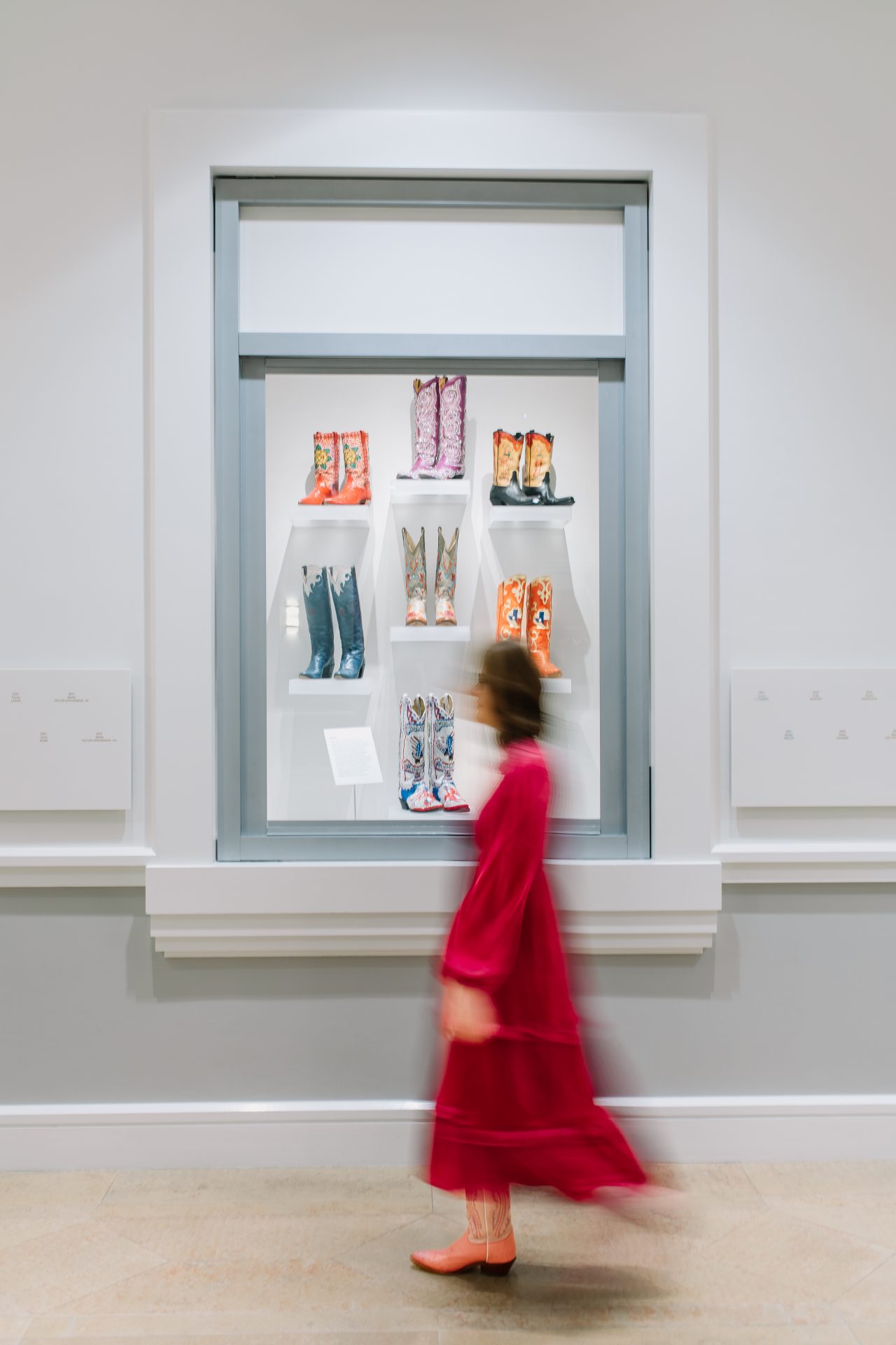 A person in a red dress walks past a display case with colorful cowboy boots on shelves in a gallery setting.