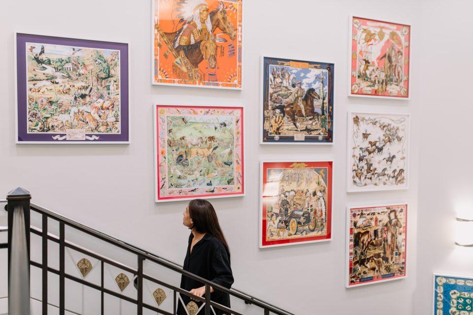 A woman walks up a staircase next to a wall displaying nine colorful, framed artworks depicting various scenes and animals. The wall is white and the staircase has black railings.