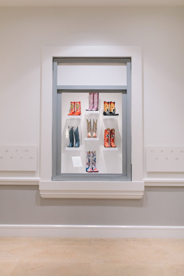 A museum display case featuring eight pairs of colorful, patterned cowboy boots arranged on three shelves against a white background.