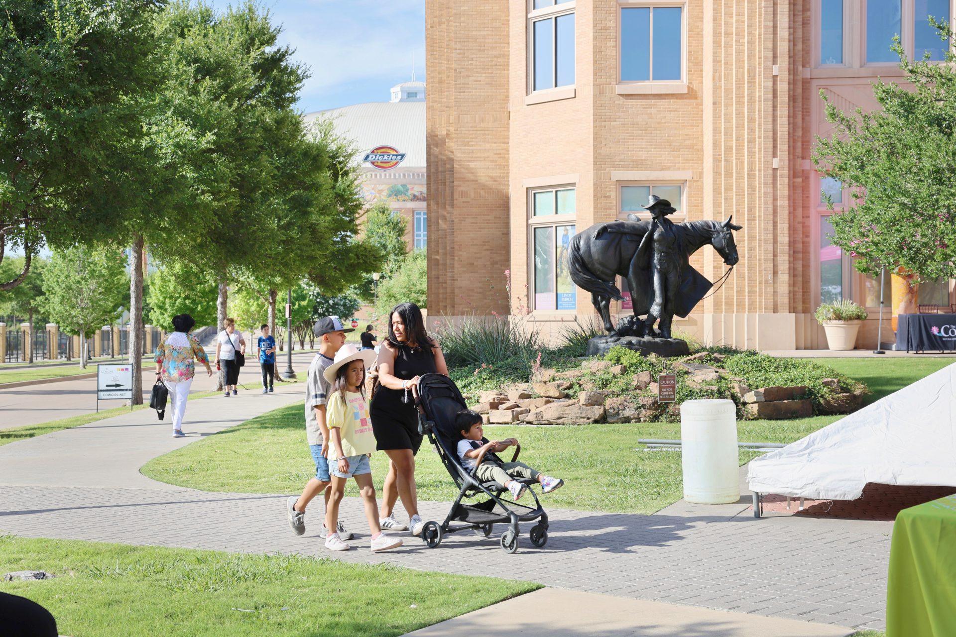 A family with two kids and a stroller walks on a sidewalk near a building with a cowboy statue on a horse. Other people are in the background on a sunny day.