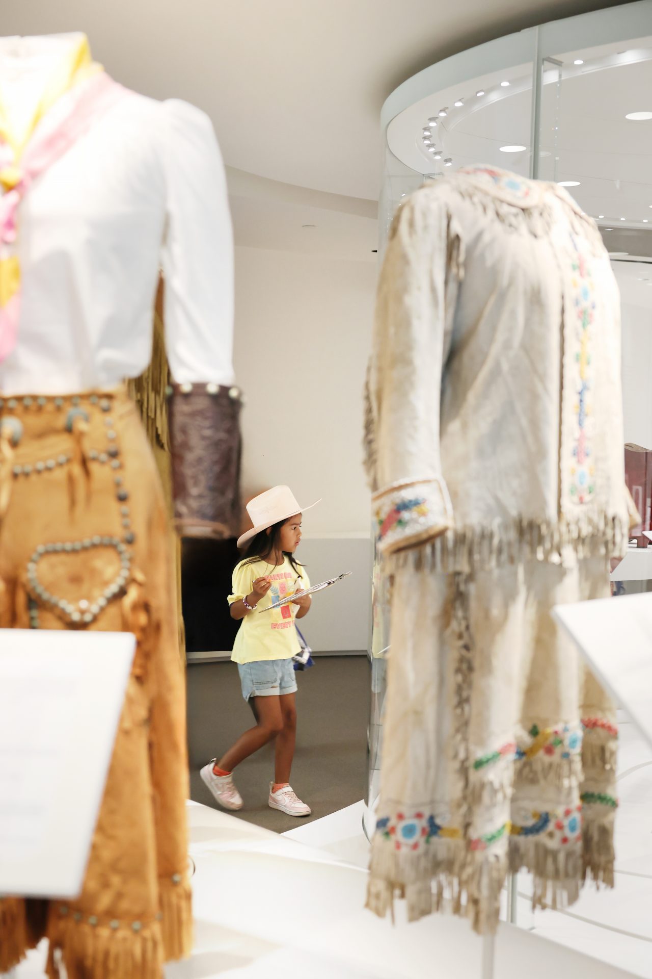 A young girl in a pink hat walks through a museum exhibit featuring traditional clothing with fringe and beadwork, looking at a display while holding a notepad.