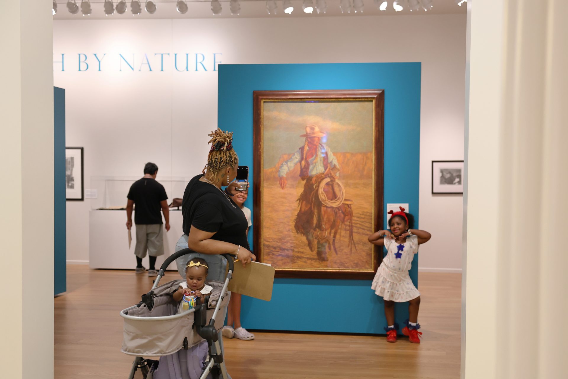 A woman with a stroller takes a photo of a smiling girl posing in front of a cowboy painting at an art museum. Other visitors view artwork in the background.