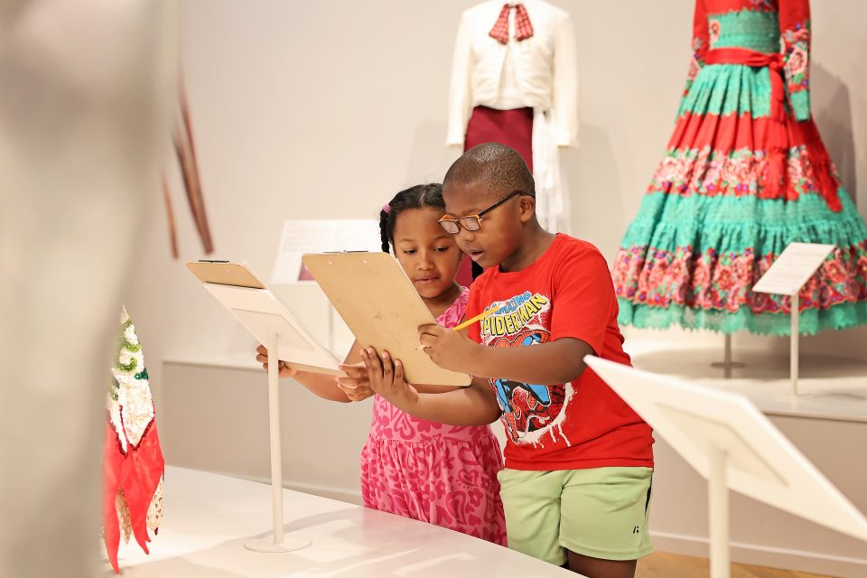 Two children stand in a museum, reading from clipboards. Behind them, colorful dresses are displayed. The children appear engaged and focused on their activity.