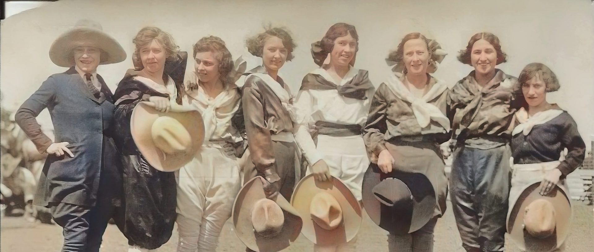 Eight women in early 20th-century attire stand side by side outdoors, smiling and holding large hats. The women wear matching outfits with sashes, and the wind is blowing their hair. The scene appears to be from a historical event.