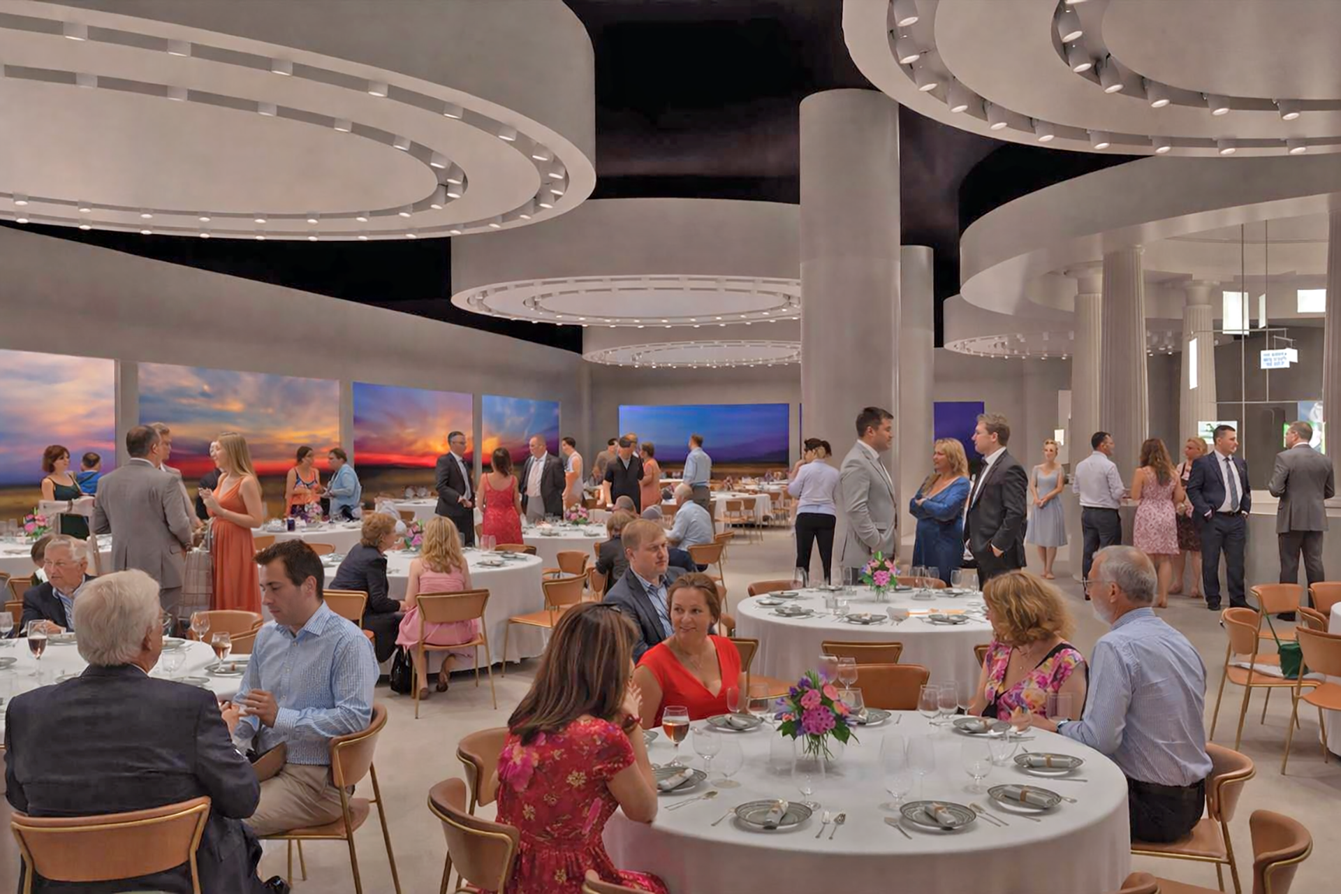 People in formal attire sit at round tables and stand mingling in a modern, elegant banquet hall with circular ceiling lights and large landscape photos on the walls. Tables have floral centerpieces and set dining places.