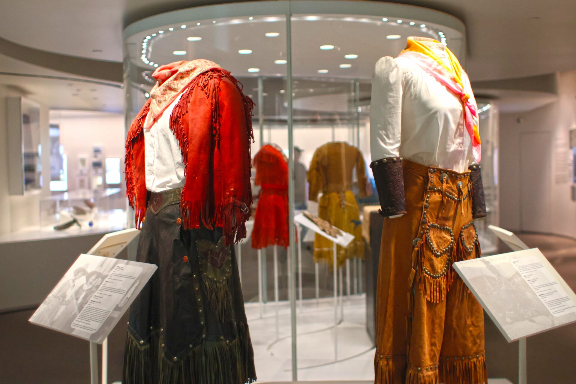 Two traditional Western outfits are displayed on mannequins in a museum exhibit, with information plaques in front and more clothing displayed in a glass case in the background.