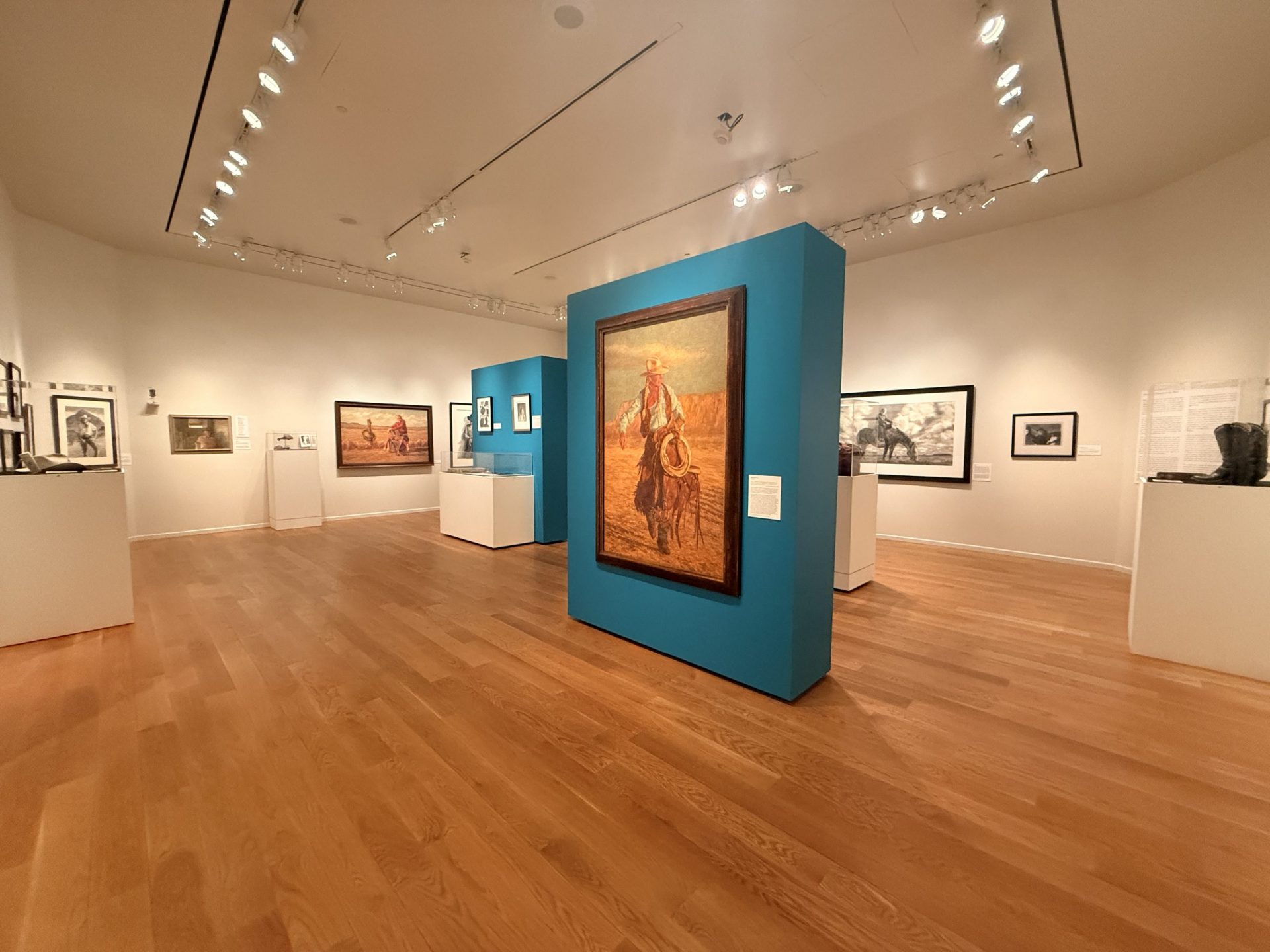 Art gallery with wooden floors, paintings on blue and white walls, sculptures on white stands, and track lighting illuminating the artworks in a spacious, modern room.
