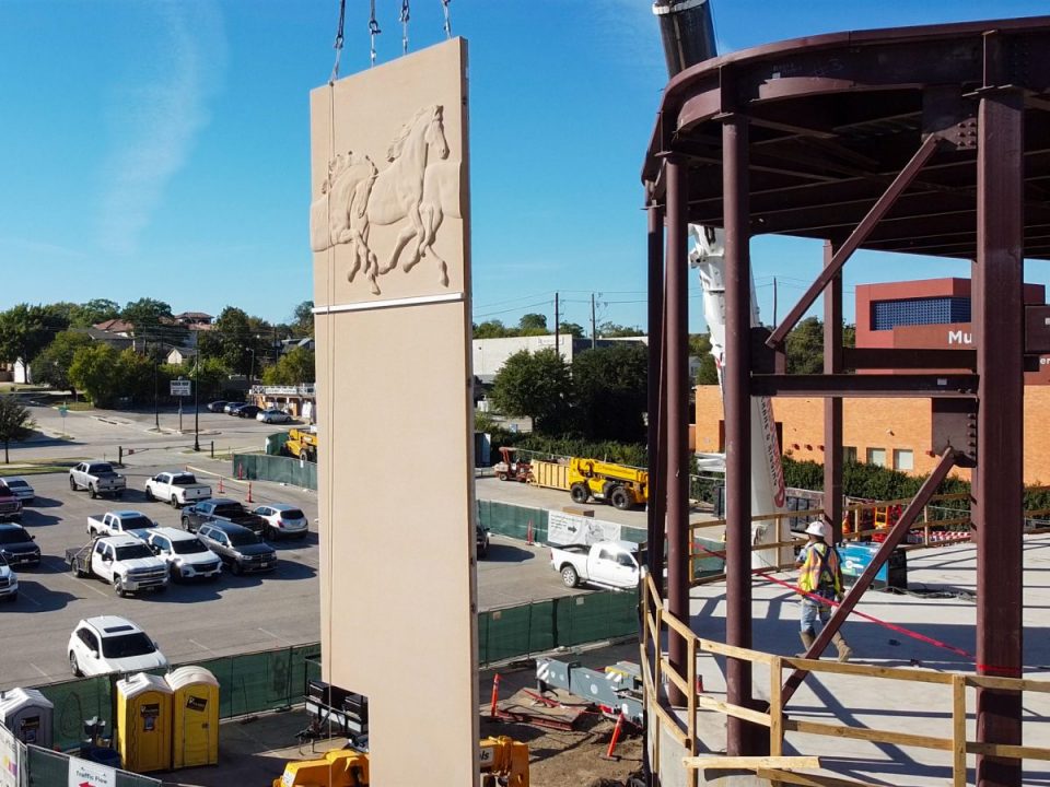 A crane lifts a tall rectangular panel with horses carved on it at a construction site, with workers nearby and cars parked in the background.
