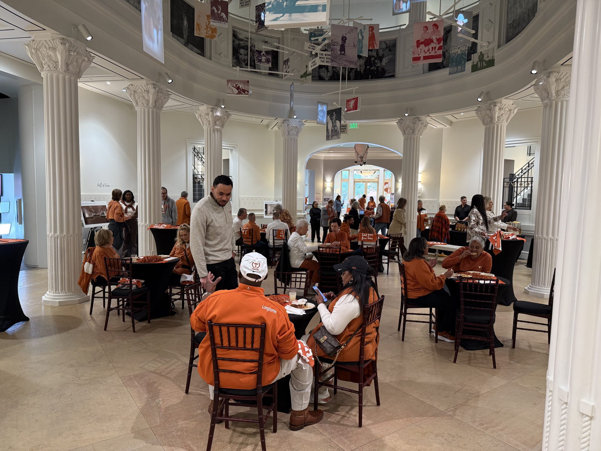 People in orange and white sit at tables in a large, bright hall with white columns. Several people are talking or using phones, and banners hang from the ceiling. The atmosphere is casual and social.