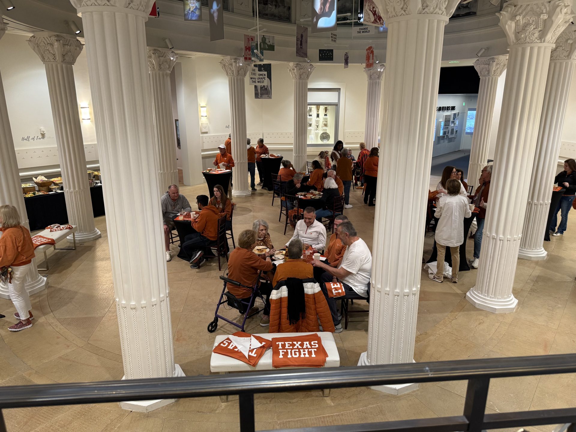 People gather around tables in a grand hall with white columns, many wearing burnt orange. A table at the front displays a Texas Fight sign, suggesting a University of Texas event. Food and drinks are visible on tables.