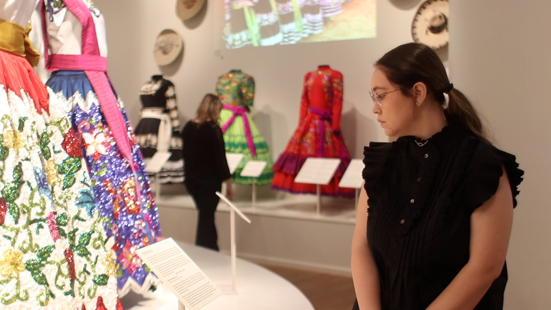 A woman in black clothing observes colorful, traditional dresses displayed in a museum exhibition with descriptive placards and projected images in the background.