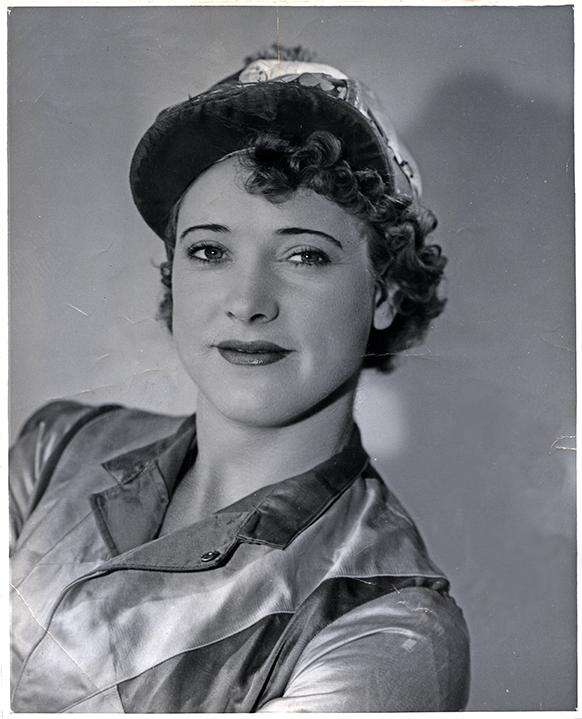 Black-and-white portrait of a smiling woman with curled hair, wearing a satin blouse and a newsboy-style hat, looking at the camera.