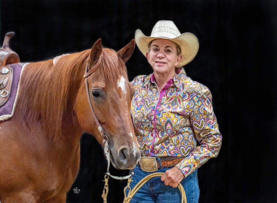Smiling cowgirl in a colorful paisley shirt and cowboy hat beside a chestnut horse against a dark background with reins in hand.