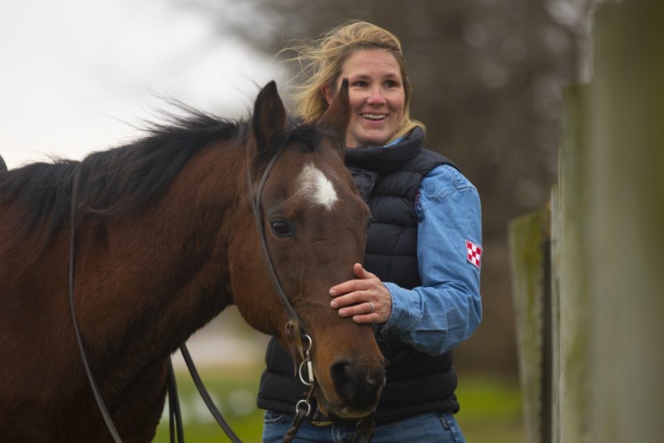 Smiling woman in a blue denim shirt and black puffer vest holding the reins of a brown horse outdoors.