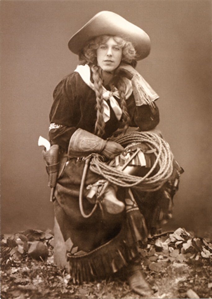 Person dressed as a cowboy with a wide hat and braided hair, holding a coiled rope and standing among leaves.