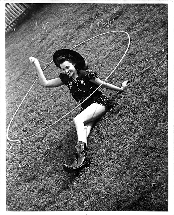 Girl in a western outfit and hat lying on grass while spinning a large hula hoop around her body, smiling at the camera.