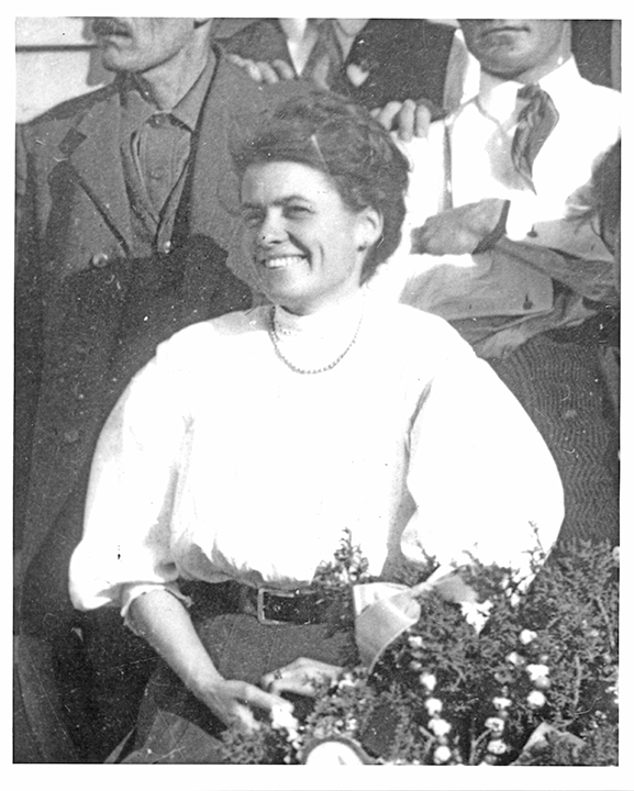 Smiling woman in a white blouse standing among a group, with flowers in the foreground.