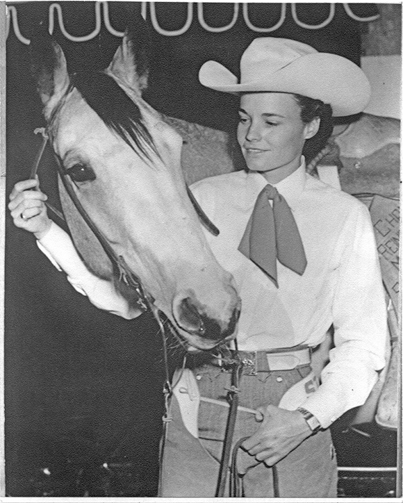 A woman wearing a white cowboy hat and neck scarf leads a light horse by the reins indoors, smiling at the animal.