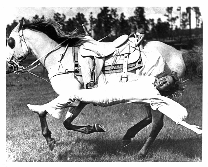 Person in white clothing is thrown from a galloping white horse during a rodeo, lying on their side as the horse bucks nearby