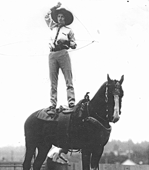 Cowboy in a wide-brimmed hat stands on the saddle of a horse, twirling a lasso overhead.