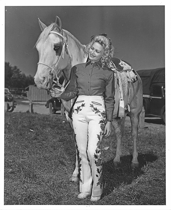 Woman in western riding outfit with embroidered white pants stands beside a saddled horse, smiling and holding its bridle.