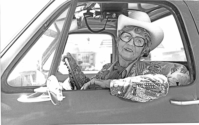 Elderly woman wearing a cowboy hat and glasses sits in the driver's seat of a vintage pickup, smiling.