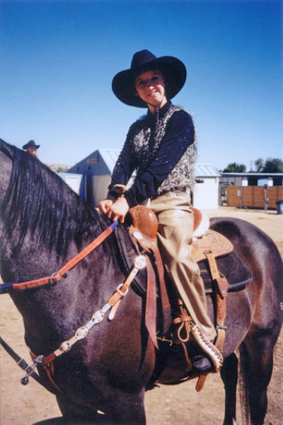 Person wearing a wide-brimmed hat rides a dark horse equipped with Western tack in an outdoor arena, smiling at the camera.